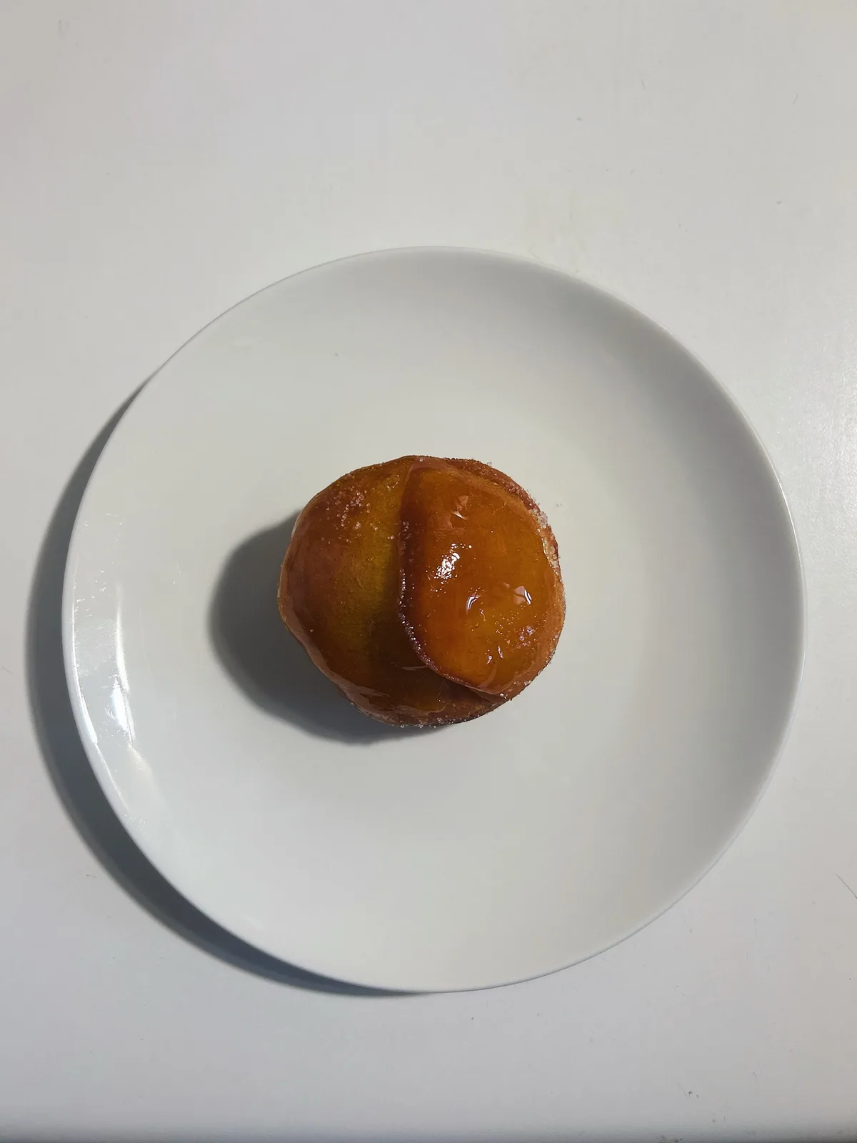 A single, round, glazed donut sits centered on a plain white plate against a white background. The donut has a shiny…