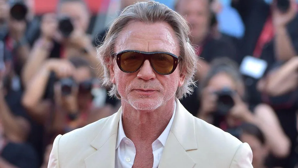 A man with gray hair and sunglasses, wearing a cream-colored suit jacket over a white shirt, stands in front of a blurred…