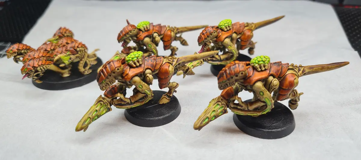 Five hand-painted fantasy miniatures, resembling armored, reptilian creatures with orange and green coloration, are posed on…