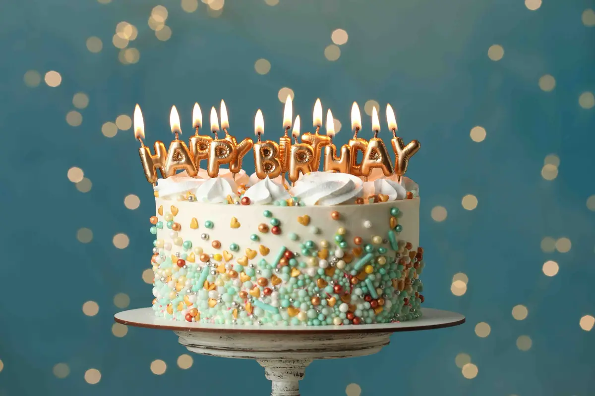 A festive birthday cake with white frosting, colorful sprinkles, and gold letter candles spelling 'HAPPY BIRTHDAY' that are…