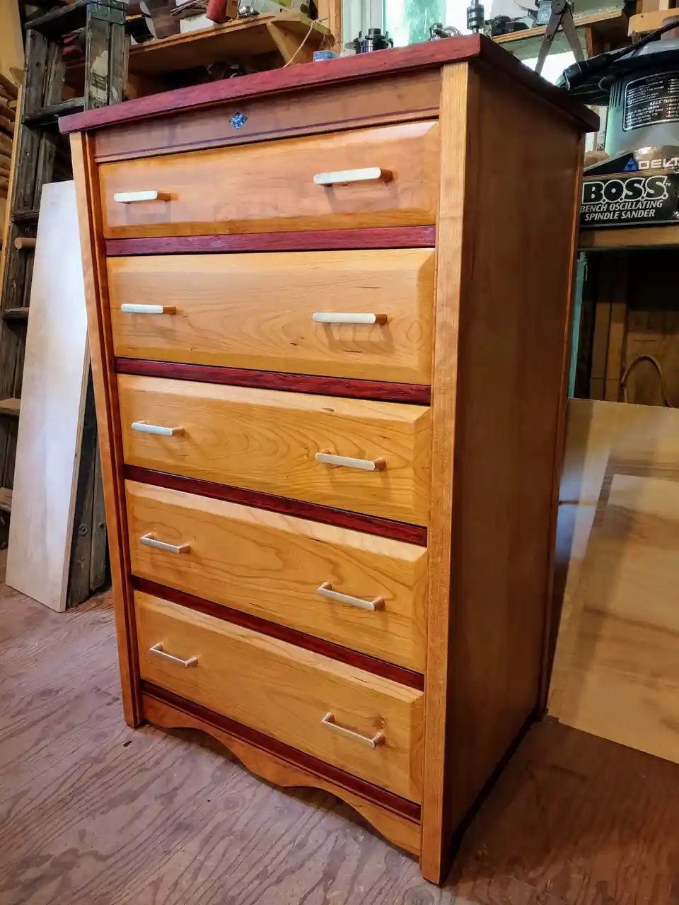 A tall, custom-made wooden dresser with five drawers stands in a cluttered woodworking shop, featuring light-colored wood…