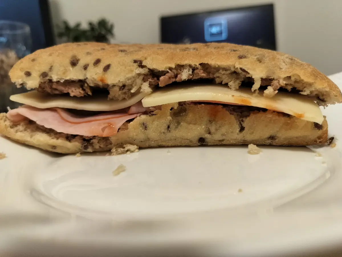 A close-up photo shows a partially eaten sandwich on a white plate, with visible layers of ham, cheese, and meat inside a…