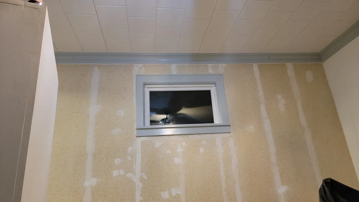 A room under renovation with drywall mud applied in vertical stripes on the walls, a small window with a grey frame, and a…