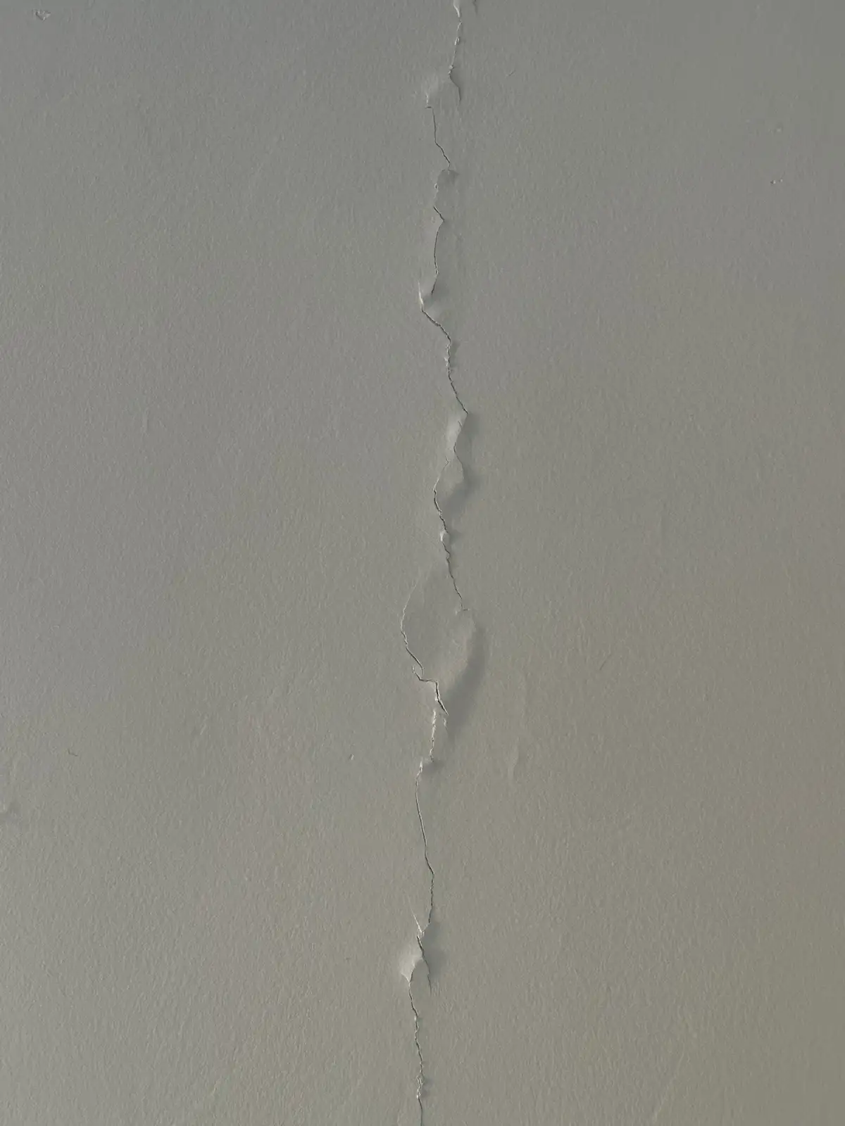 A close-up photo shows a long, jagged crack running vertically down a plain, light gray wall. The crack is uneven and…
