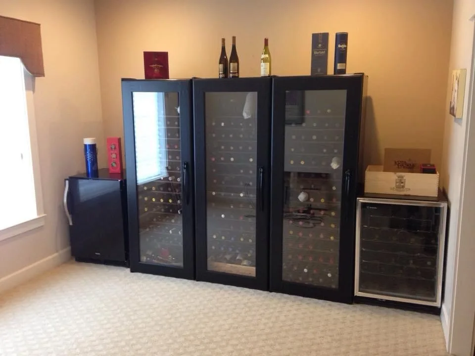 A room with three large black wine refrigerators lined up, each with glass doors showing rows of wine bottles inside. On top…