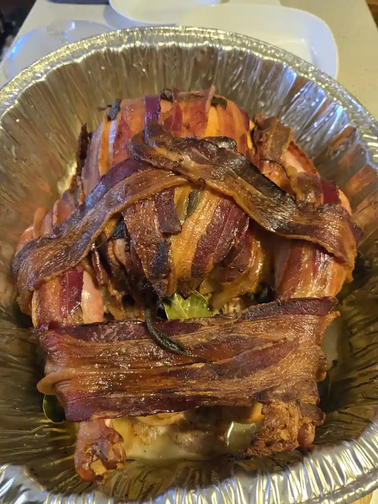 A close-up photo shows a roasted turkey breast heavily wrapped in crispy, browned bacon strips, sitting in a shiny aluminum…