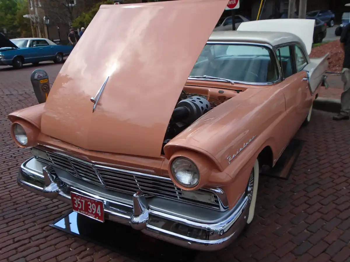 A vintage 1957 pink Lincoln Continental with its hood open, revealing a large, black, perforated gun barrel instead of an…