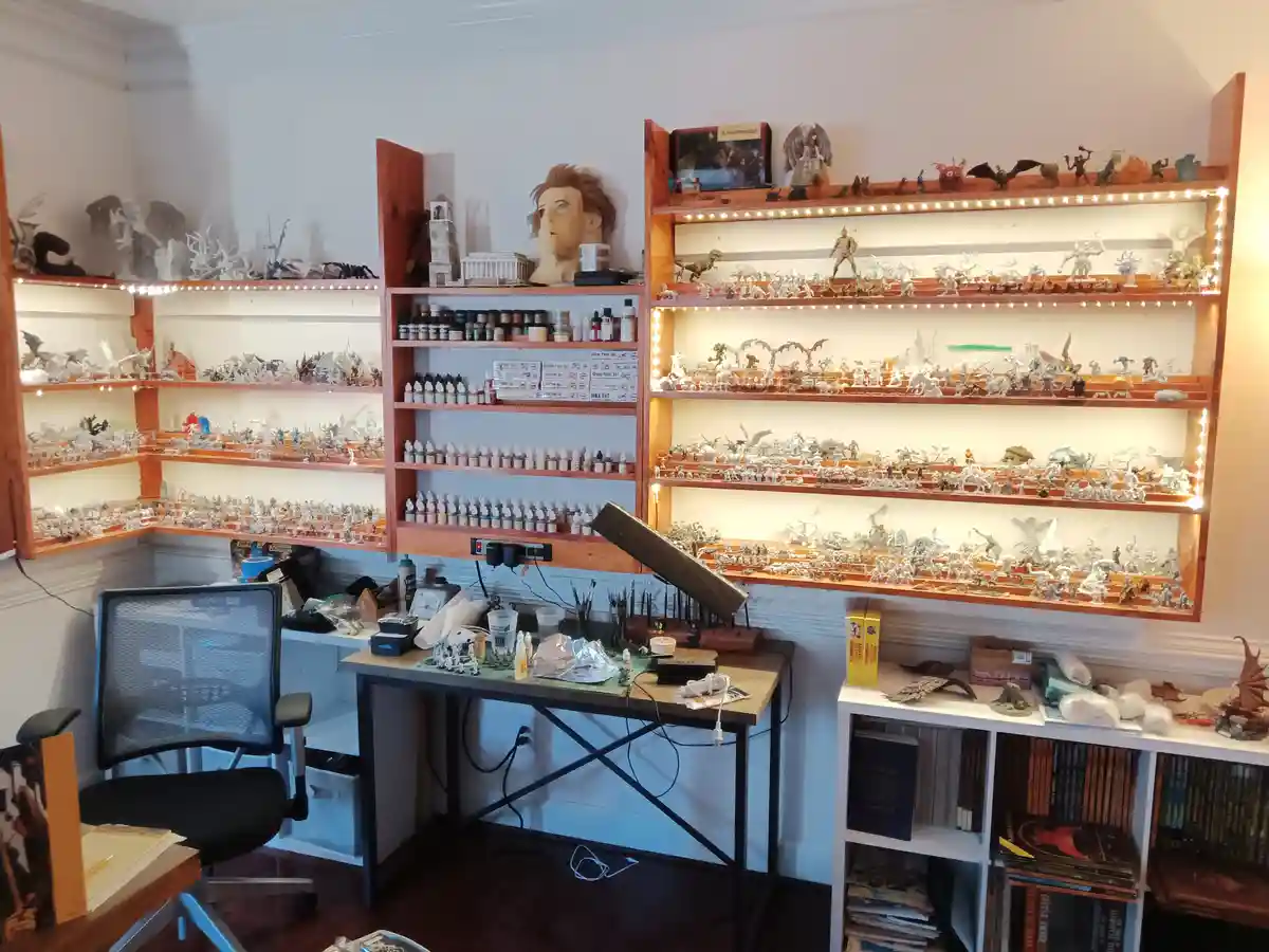 A room filled with wooden shelves displaying hundreds of unpainted fantasy miniatures, with paint bottles on a central shelf…
