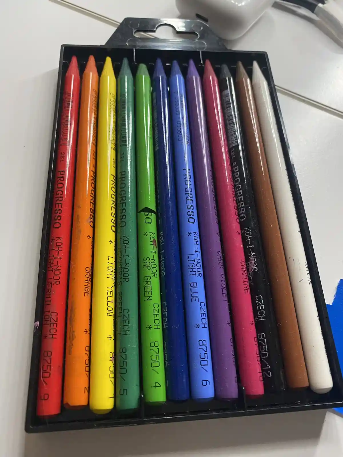 A close-up photo shows a black plastic tray holding a set of colorful Koh-I-Noor brand crayons, arranged in a rainbow order…