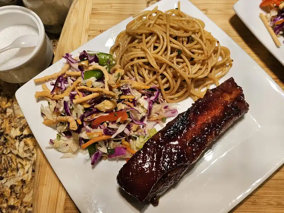 A white plate holds a meal with glossy barbecue ribs, stir-fried noodles, and a colorful coleslaw with purple cabbage…