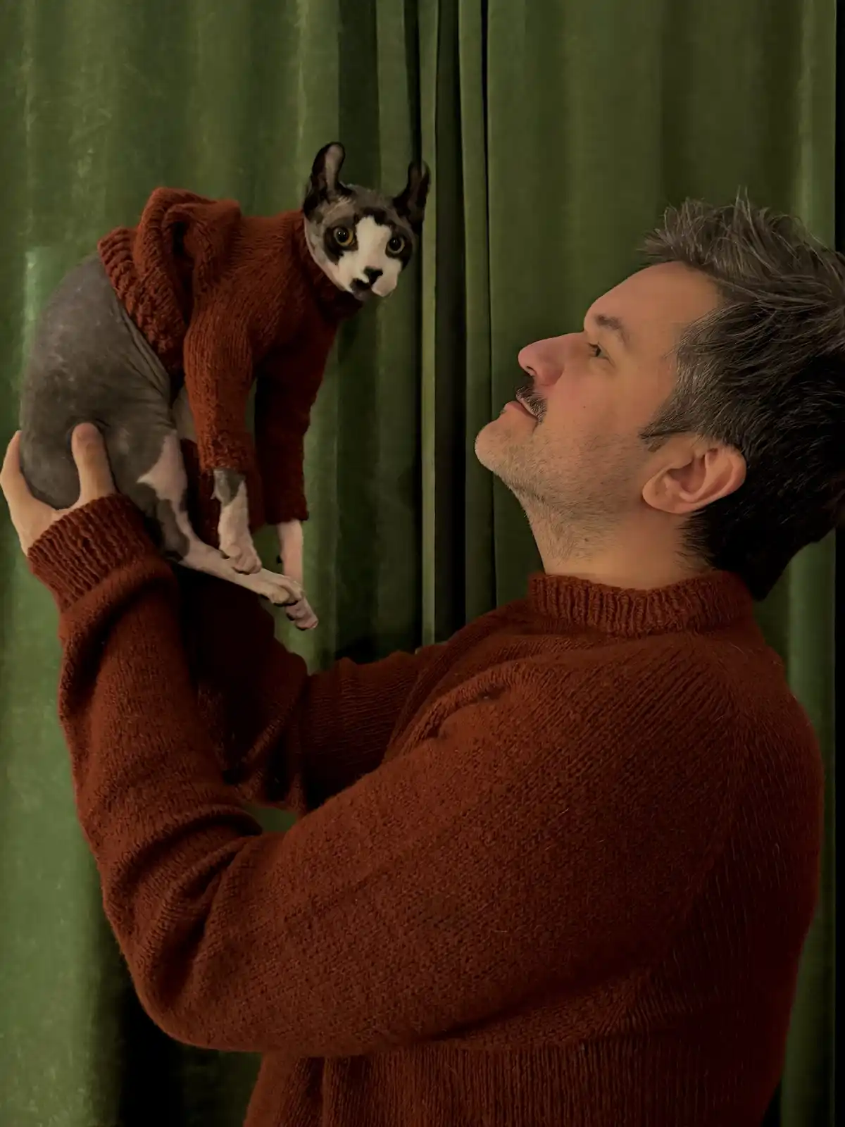 A man in a rust-colored turtleneck sweater holds up a hairless cat wearing a matching rust-colored sweater against a green…