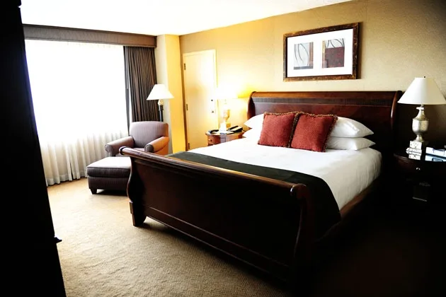 a well-lit, spacious hotel room with a large wooden sleigh bed, two nightstands with lamps, a chair and ottoman near a…
