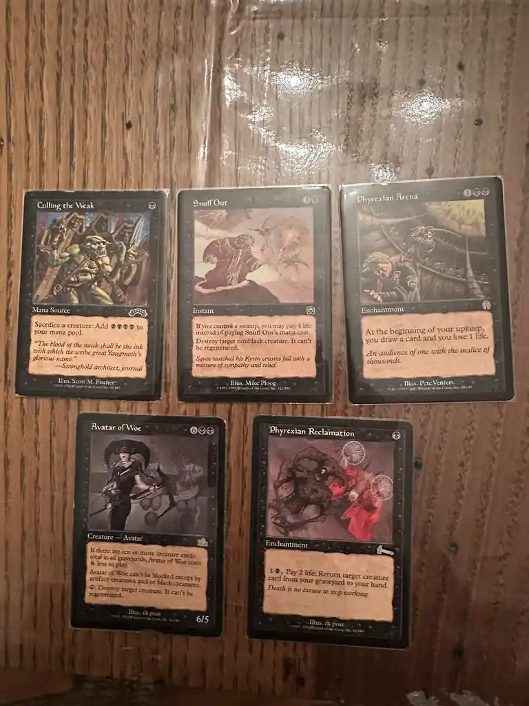 Five Magic: The Gathering cards are laid out on a wooden surface. The cards are 'Calling the Weak', 'Snuff Out', 'Phyrexian…