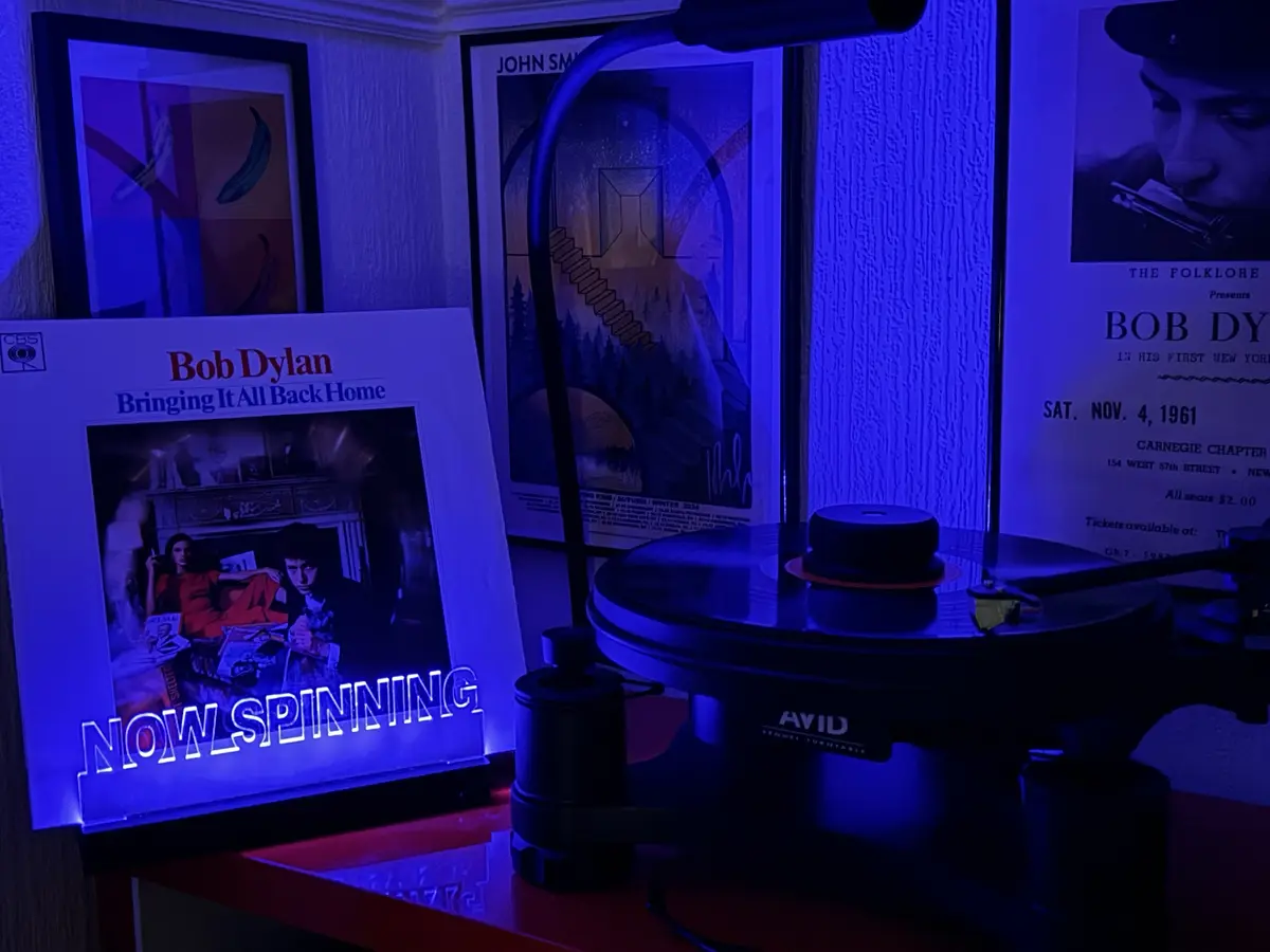 A vinyl record player sits on a red surface under blue lighting, next to a glowing sign displaying the album cover of Bob…