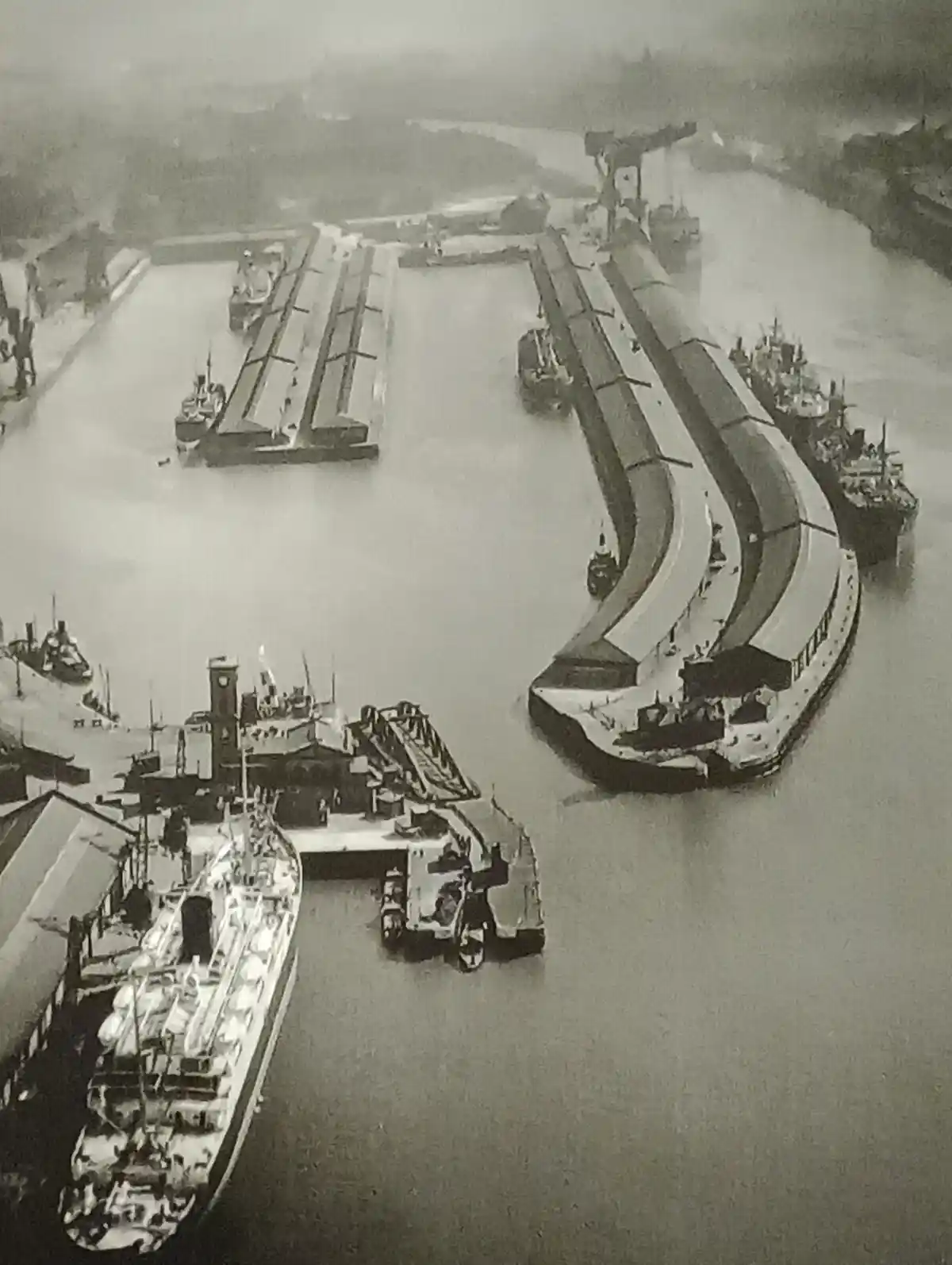 A black-and-white aerial photograph of a busy, historic harbor with multiple ships docked at curved and straight piers…
