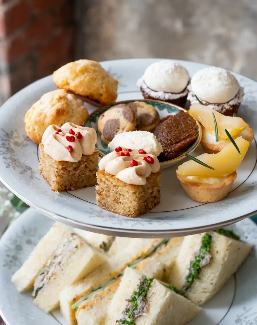 A two-tiered serving tray holds an assortment of delicate pastries and sandwiches. The top tier features small cakes with…