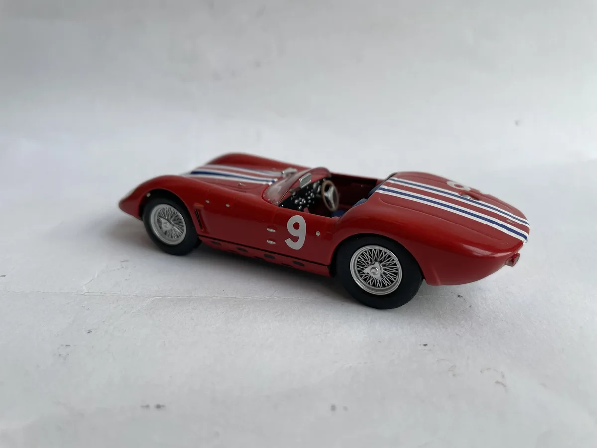 A red die-cast model of a vintage race car with white and blue stripes, number 9 on its side, sits on a plain white surface…
