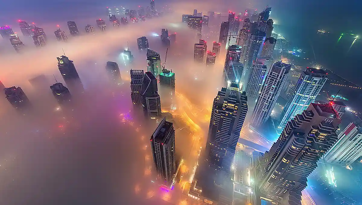 An aerial nighttime photo of a futuristic cityscape with skyscrapers emerging from thick, colorful fog, illuminated by…