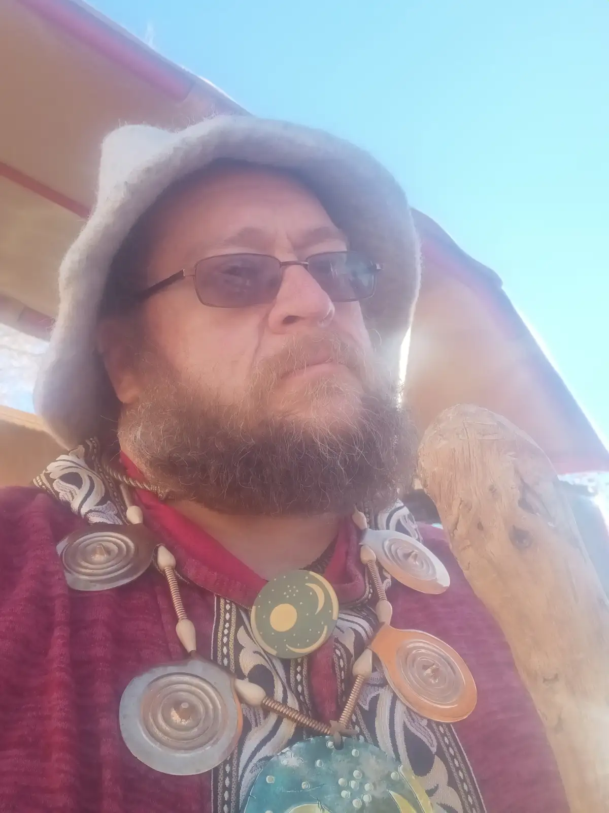 A man with a full beard, glasses, and a grey hat looks upward with a serious expression, wearing a red shirt and an…