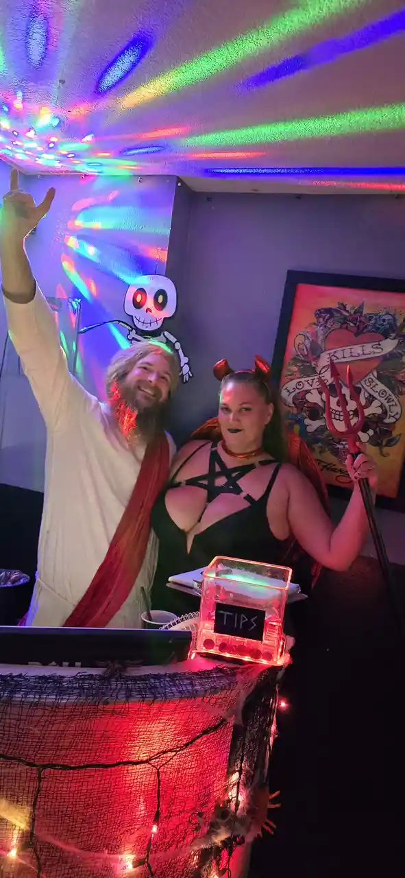 Two people dressed as Jesus and the devil stand behind a decorated counter with a glowing 'TIPS' box. The Jesus figure wears…