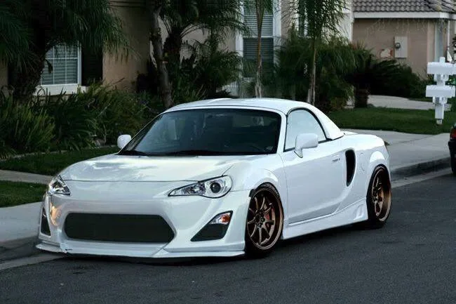 A white modified sports car, likely a Scion FR-S or similar, is parked on a residential street with palm trees and houses in…