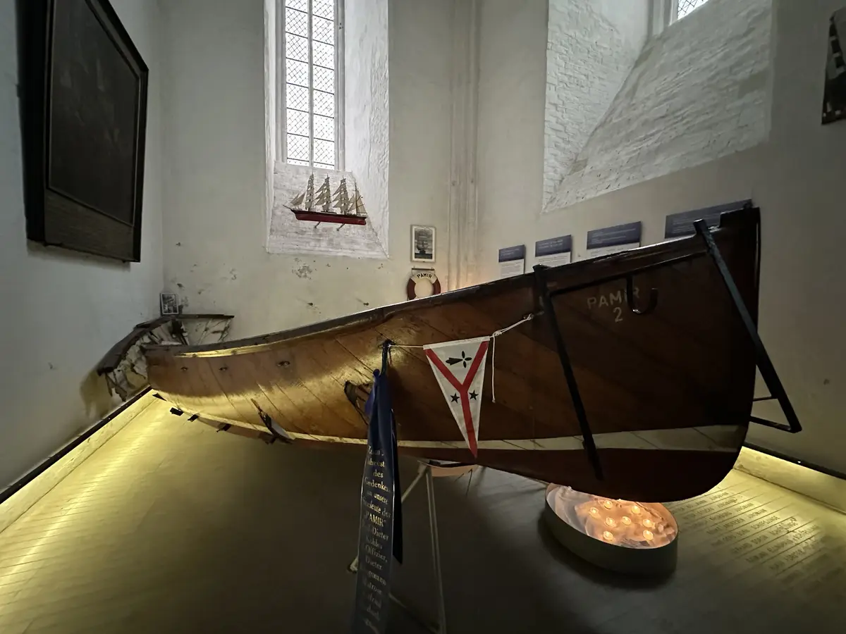A wooden lifeboat labeled 'PAMIR 2' is displayed indoors on a stand, illuminated by lights beneath it. A small flag and a…