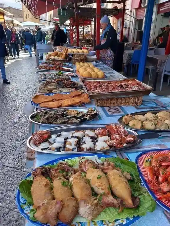 A long outdoor market stall displays an abundant variety of cooked Mediterranean foods including fried squid, grilled…