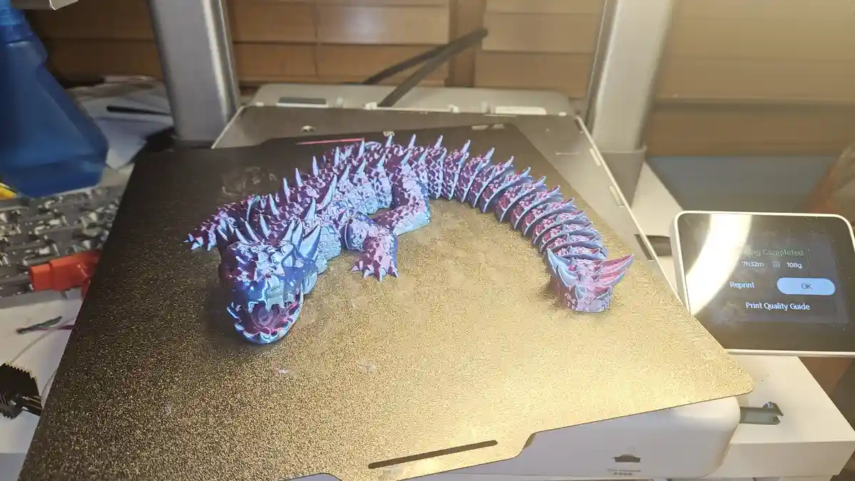 A 3D-printed dragon model with a gradient of blue and purple sits on the build plate of a 3D printer. The printer's…