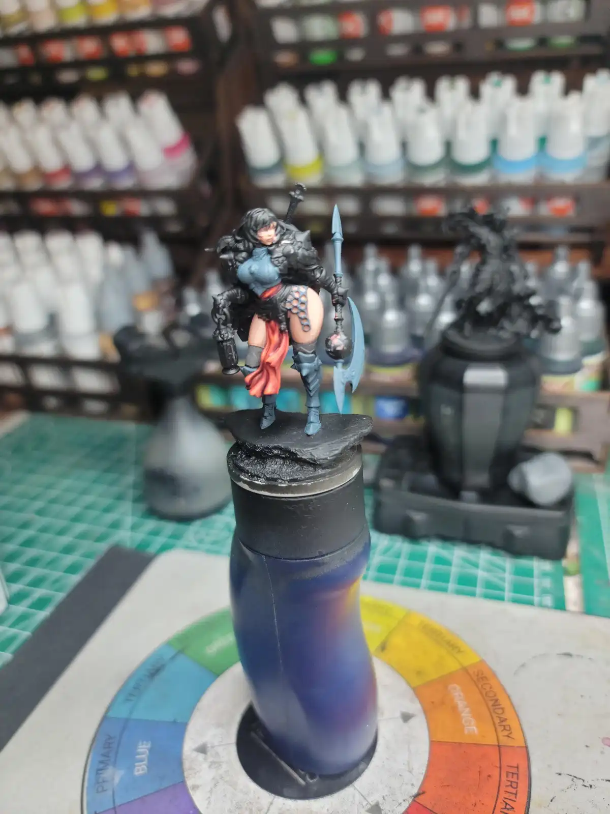 A painted fantasy miniature warrior figure, holding a spear and shield, stands atop a blue and purple gradient paint bottle…