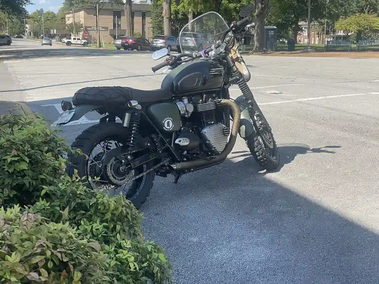 A matte olive-green Triumph motorcycle with camouflage accents is parked in an asphalt lot next to green bushes, featuring a…