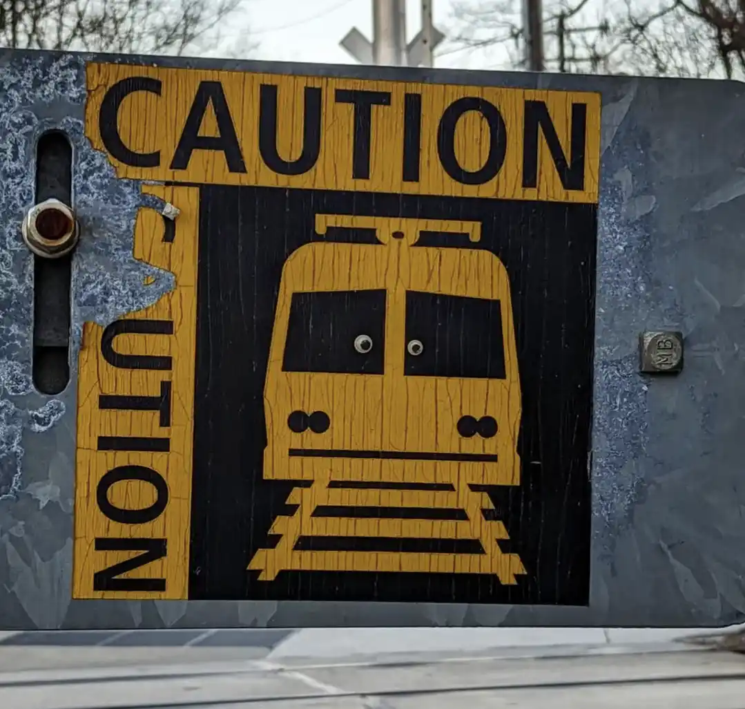 A weathered, yellow-and-black railroad crossing sign shows a train with googly eyes, and the word 'CAUTION' is visibly…