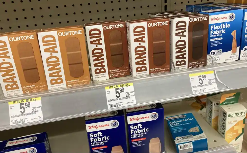 A store shelf displays boxes of Band-Aid Ourtone flexible fabric bandages in brown and white packaging, priced at $5.99…