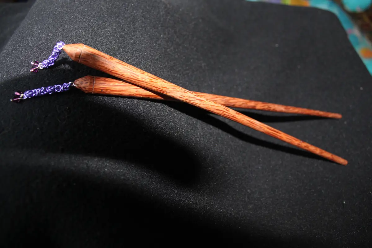 Two handcrafted wooden hair sticks with purple beaded chains lie crossed on a black fabric surface. The overlaid text reads…