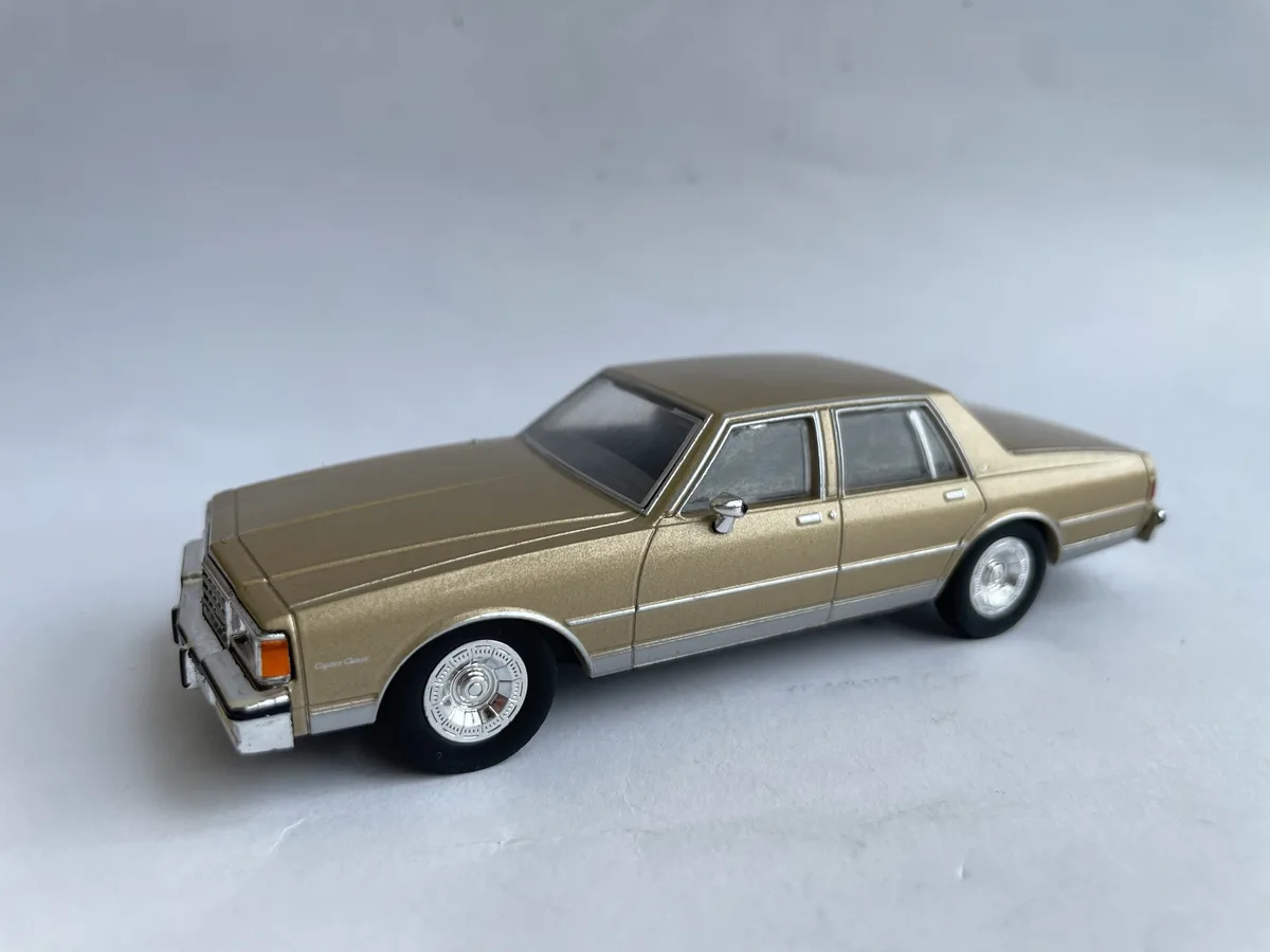 A gold-colored die-cast model of a classic American sedan, likely a Chevrolet Caprice Classic, is photographed against a…