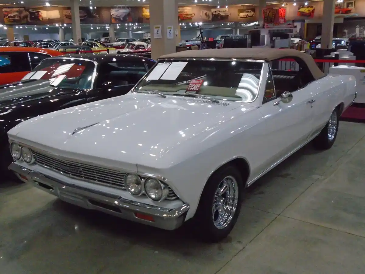 A white classic convertible car with chrome details is displayed in a museum or showroom, surrounded by other vintage cars…