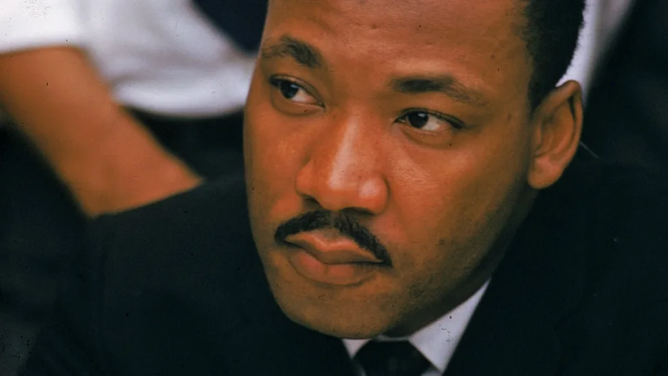 a close-up photo of Martin Luther King Jr. looking sideways with a skeptical, unimpressed expression. He is wearing a dark…
