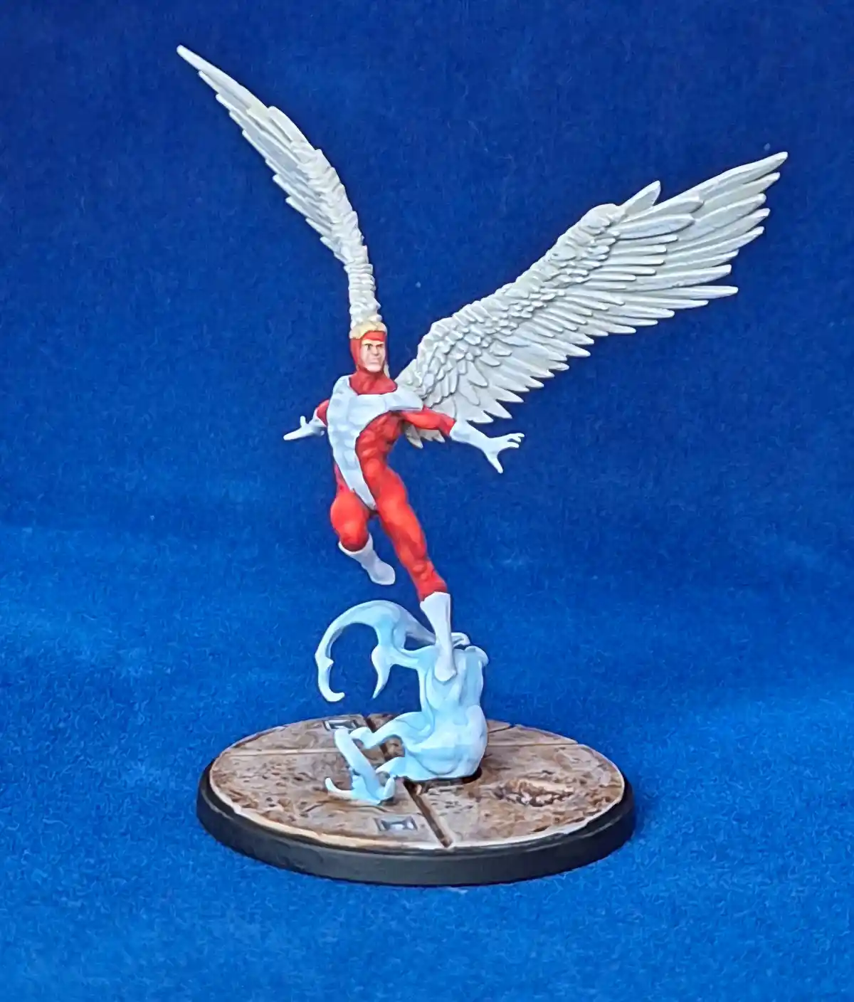 A painted miniature figurine of a red-and-white costumed angel with large white wings stands triumphantly on a blue base…