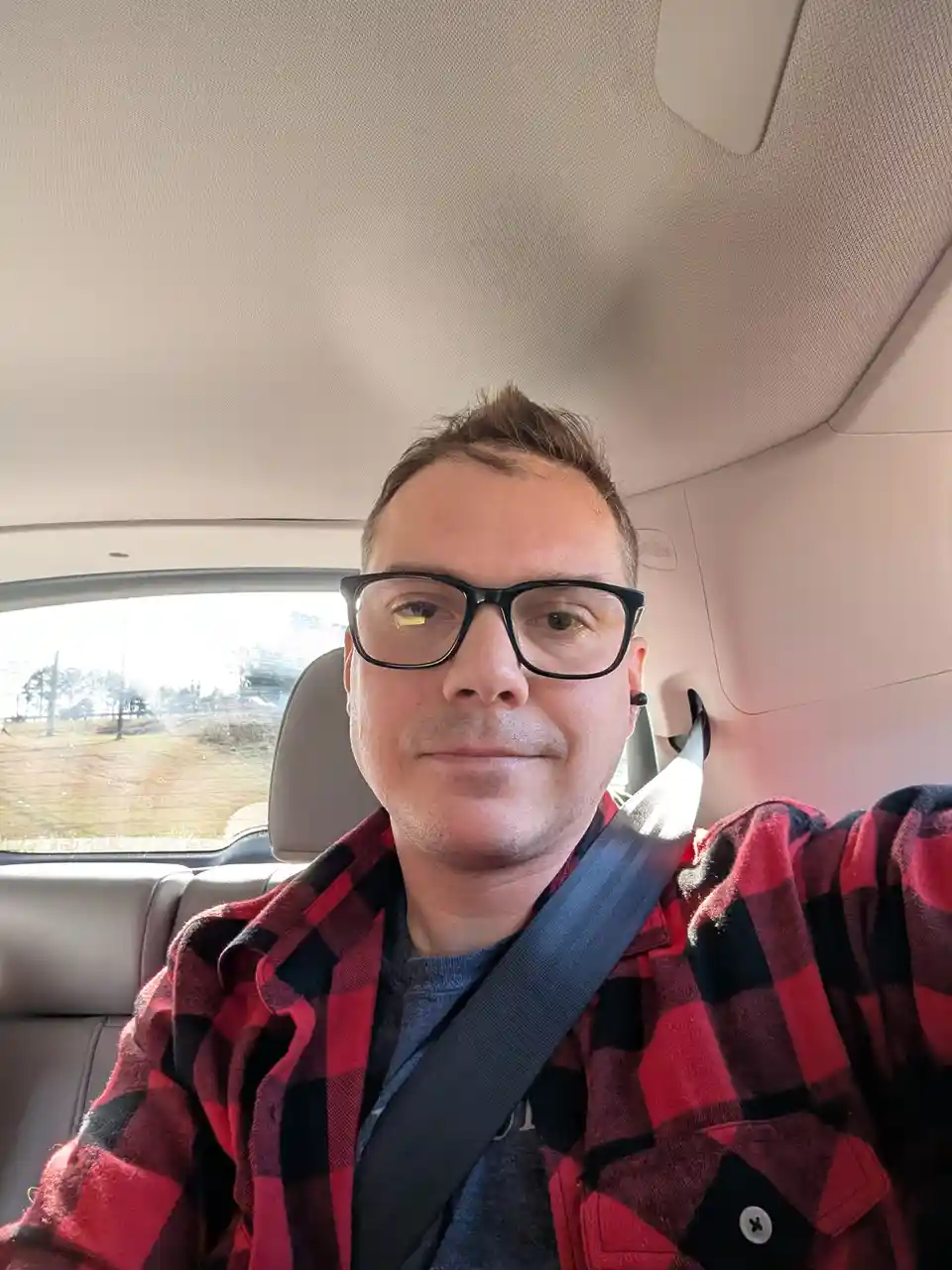 A man wearing black-rimmed glasses and a red-and-black plaid shirt sits in the driver's seat of a car, buckled in with a…