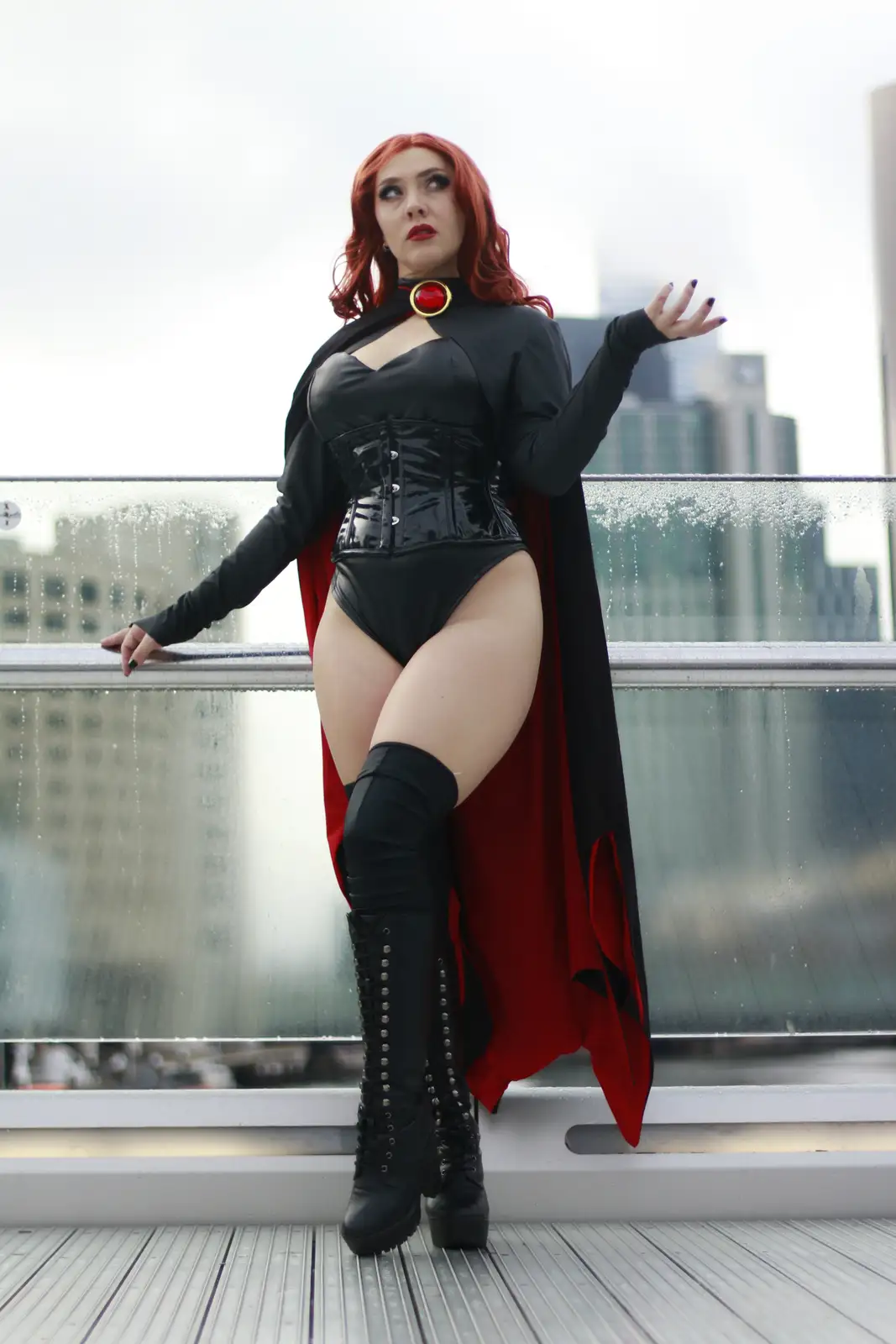 A person in a Black Widow cosplay, featuring a black corseted bodysuit, red cape, and thigh-high boots, poses dramatically…