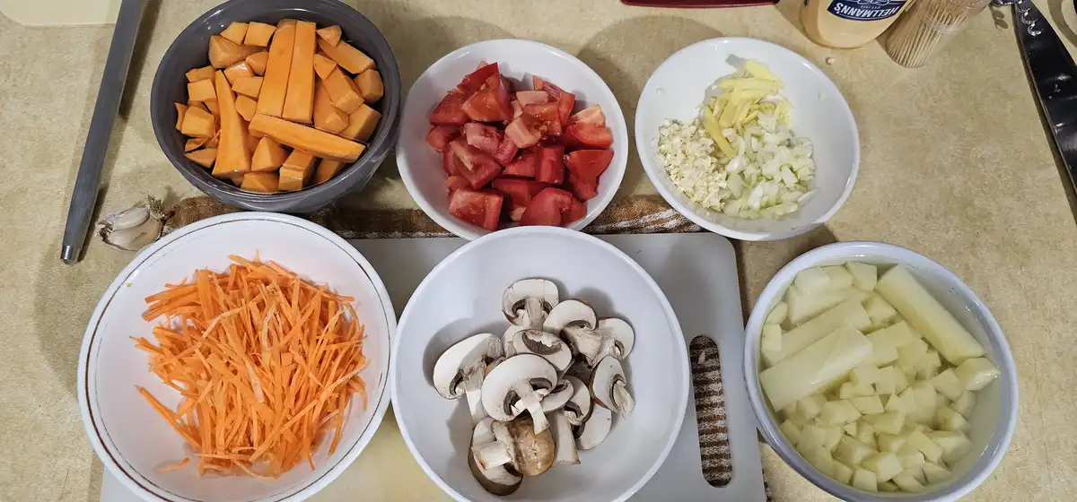 Six bowls of prepped vegetables — chopped sweet potato, tomato, garlic and ginger, shredded carrot, sliced mushrooms, and…