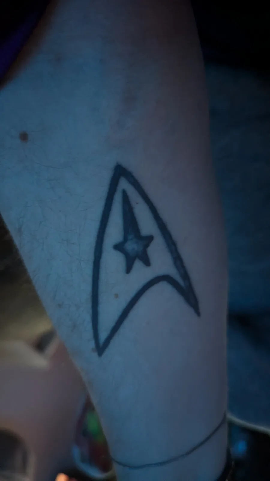 A close-up photo shows a black ink tattoo of the Starfleet insignia from Star Trek on a person's forearm. The tattoo is a…
