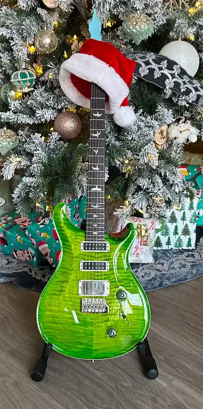 A bright green electric guitar with a Santa hat perched on its neck stands in front of a decorated Christmas tree with…