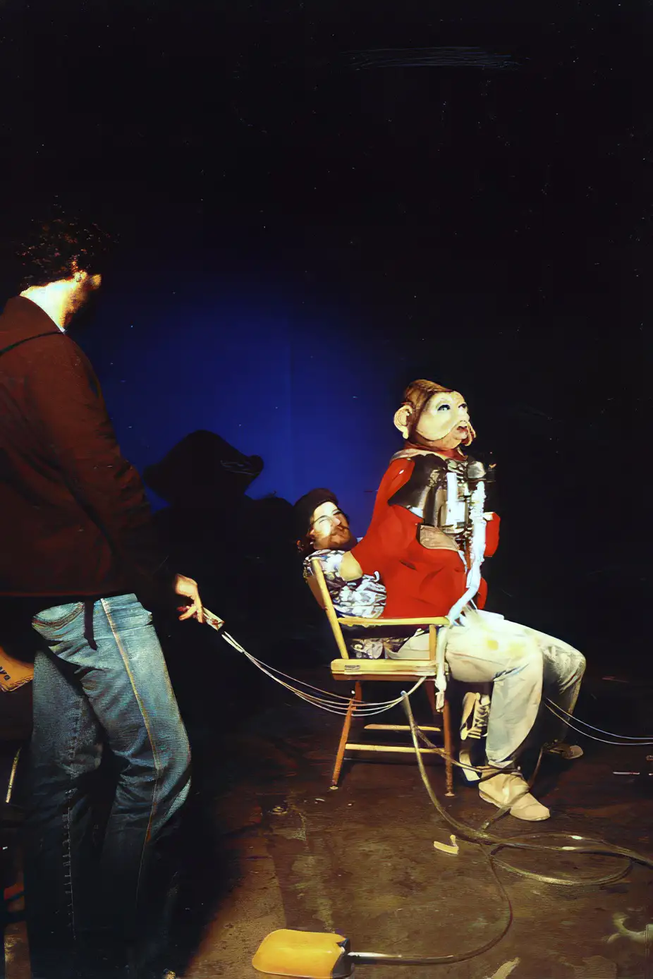 A person in a red jacket and jeans stands holding a leash attached to a person seated in a wooden chair. The seated person…