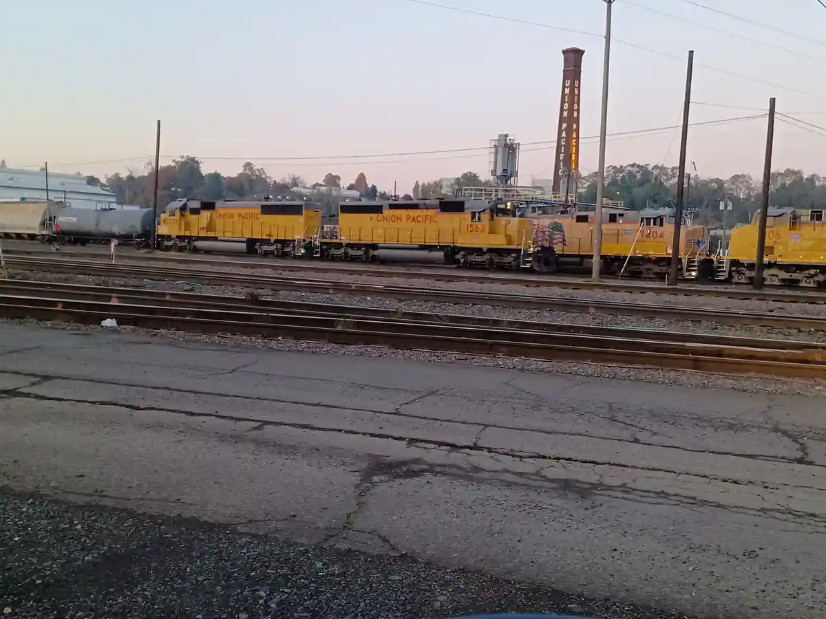 A row of yellow Union Pacific locomotives sits on multiple railroad tracks at a rail yard during dusk, with a tall brick…