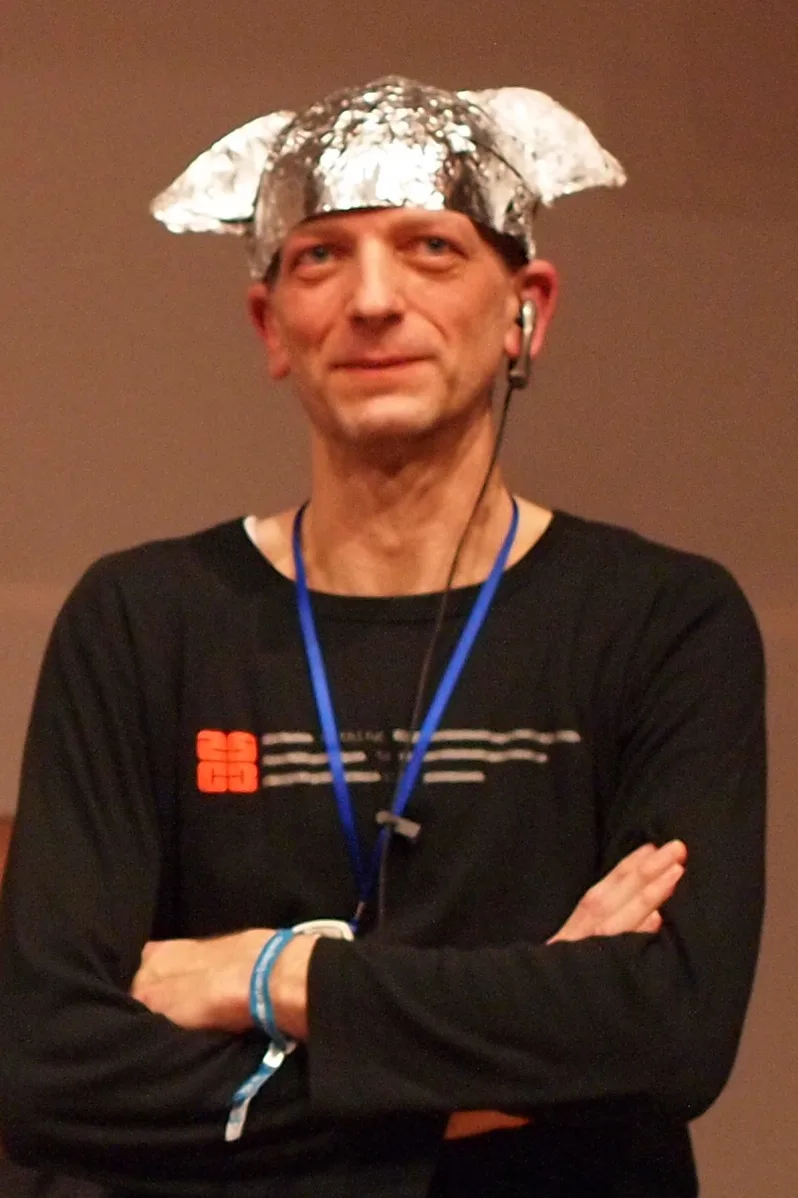 a man with arms crossed wears a black long-sleeved shirt with a blue lanyard and a silver foil hat shaped like cat ears…