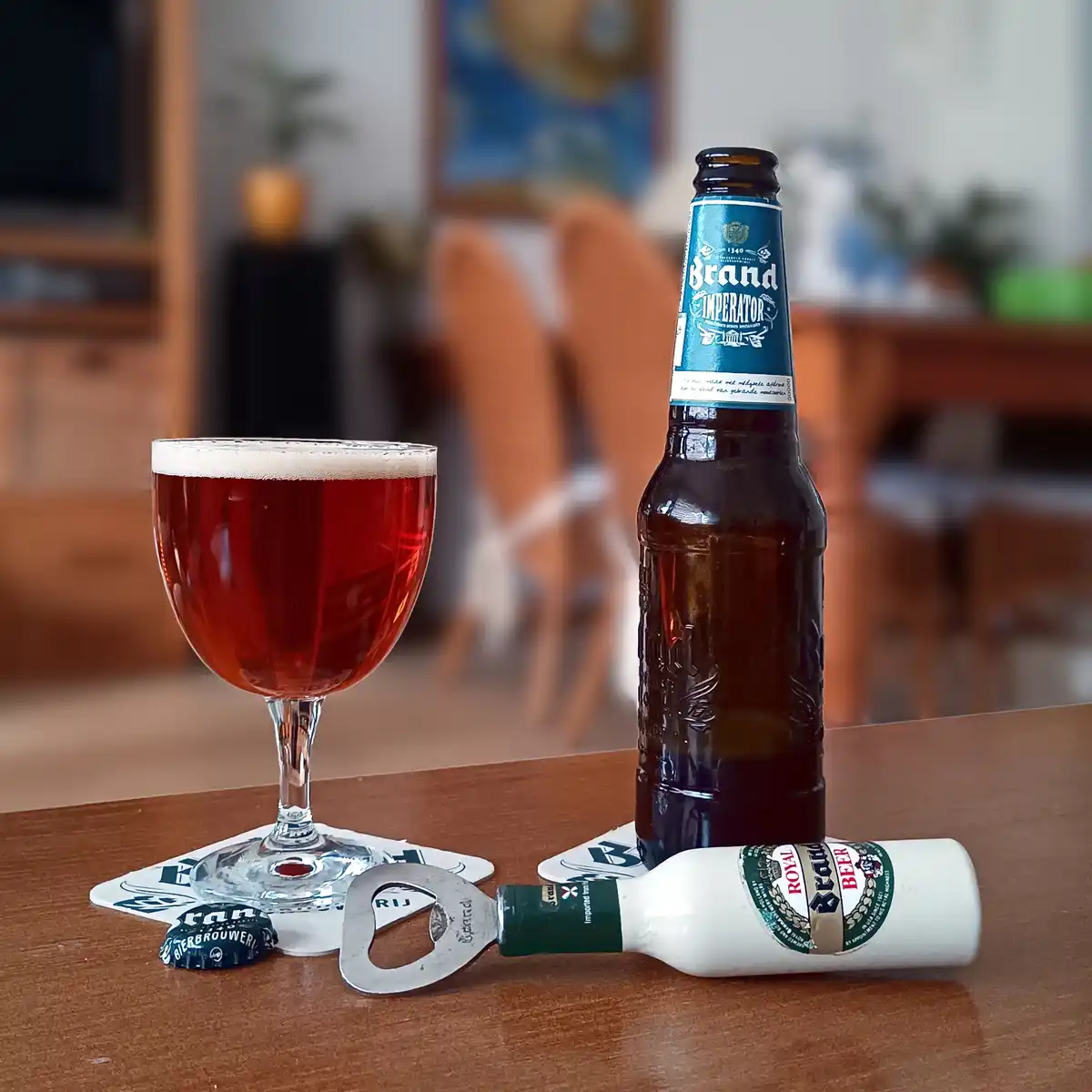 A brown glass bottle of 'Brand Imperator' beer stands next to a filled beer glass on a wooden table, with a bottle opener…