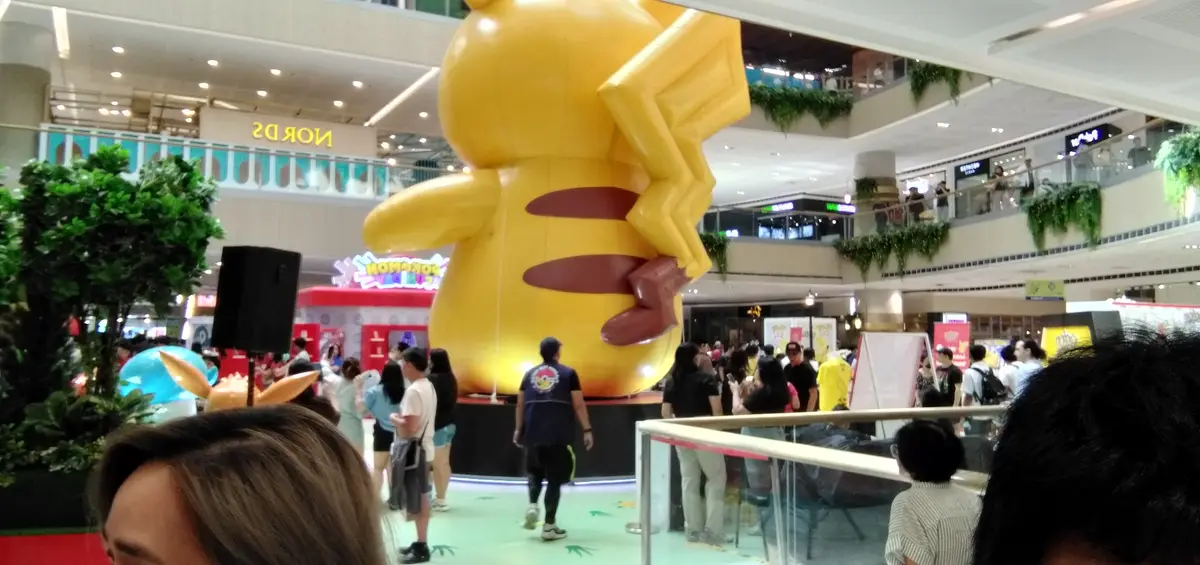 A large, yellow inflatable Pikachu statue dominates a crowded shopping mall atrium, surrounded by people taking photos and…