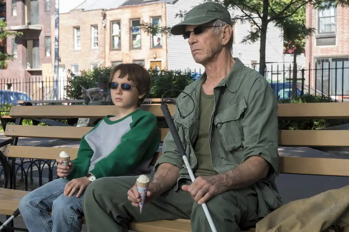 An older man with a white cane and sunglasses sits on a park bench next to a young boy also wearing sunglasses, both holding…