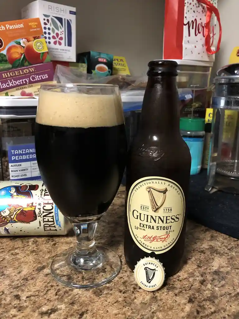 A bottle of Guinness Extra Stout beer sits next to a full glass on a kitchen counter, with the bottle cap on the counter…