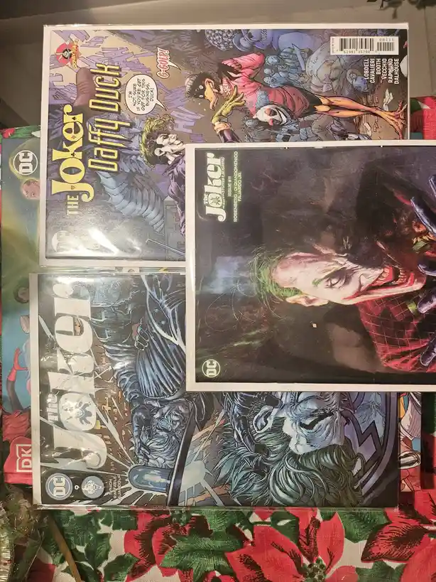 Four comic book covers featuring The Joker are arranged on a floral-patterned surface. The top-left cover shows The Joker…